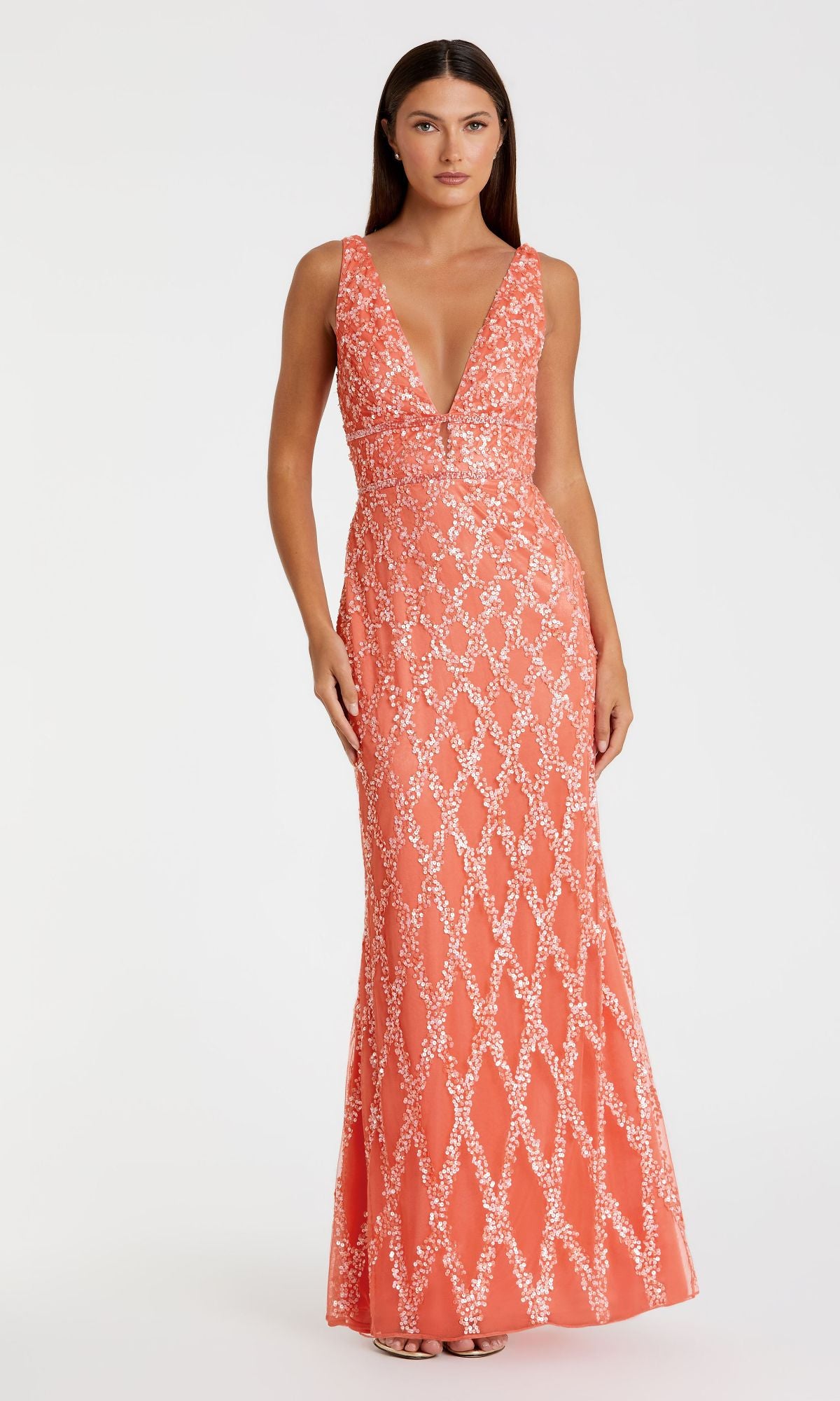 A woman models the Mac Duggal 5669 Backless Long Beaded Formal Dress, a sleeveless floor-length coral gown with a deep V-neckline, white floral patterns, and open back, standing against a plain white background.